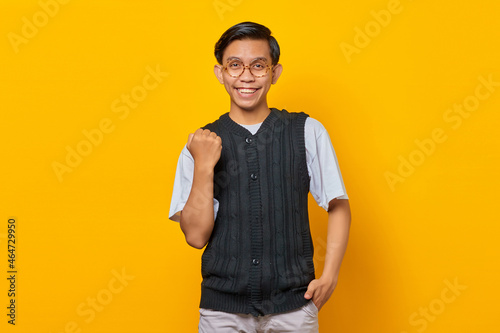 Portrait of excited young Asian man celebrating success with raised arms on yellow background