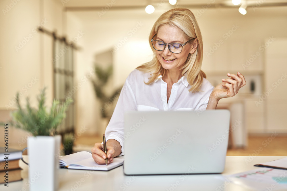 Attractive mature businesswoman smiling and making notes while having online video call using laptop, sitting at her desk in the office Attractive mature businesswoman smiling and making notes while having online video call using laptop, sitting at her desk in the office
