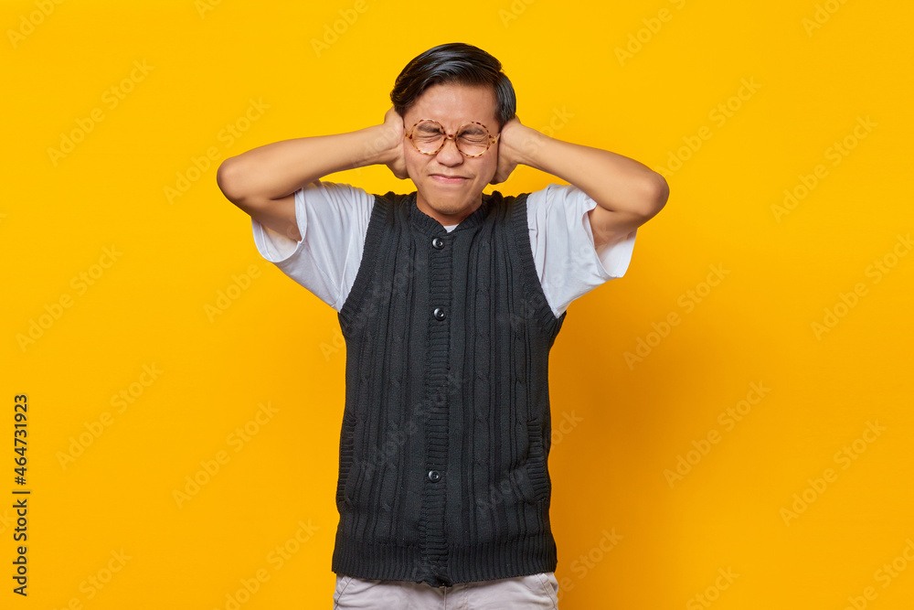 Frustrated Asian young man covering ears with hands isolated over yellow background Frustrated Asian young man covering ears with hands isolated over yellow background