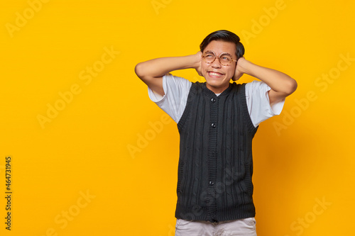 Frustrated Asian young man covering ears with hands isolated over yellow background