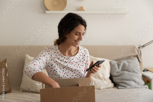 Happy consumer giving feedback to online order delivery or internet store service, using app on smartphone, opening carton box, unpacking received package at home. Shopping from home concept