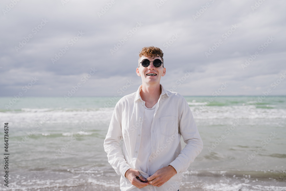 a guy in round glasses on the seashore stands with a broadly positive smile