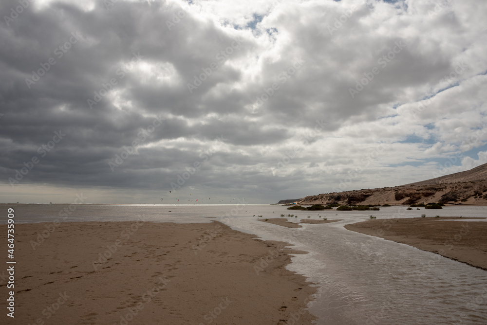 Heavy clouds over the Sotavento beach, Costa Calma, Fuerteventura, Spain. Natural brook, kitesurfers in the background. Selective focus. Heavy clouds over the Sotavento beach, Costa Calma, Fuerteventura, Spain. Natural brook, kitesurfers in the background. Selective focus.