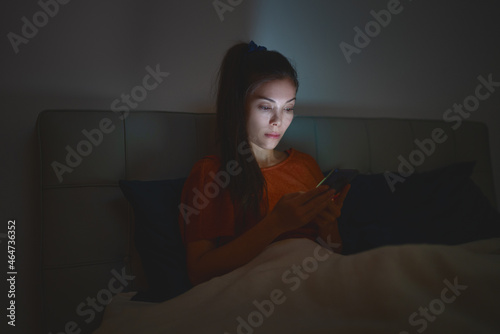 Woman sleeping using phone in bed staying up late at night reducing her sleep time which is bad for mental health and depression.