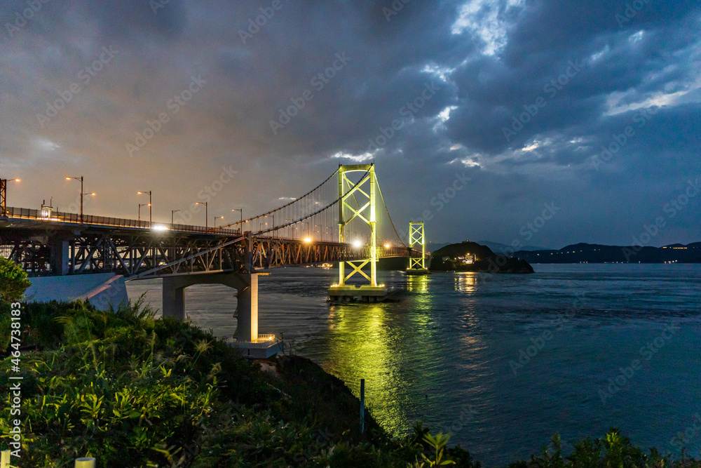 Naklejka premium Lights from suspension bridge mix with sunset glow and dramatic clouds