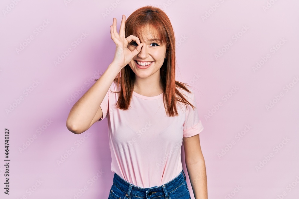 Redhead young woman wearing casual pink t shirt doing ok gesture with hand smiling, eye looking through fingers with happy face. Redhead young woman wearing casual pink t shirt doing ok gesture with hand smiling, eye looking through fingers with happy face.