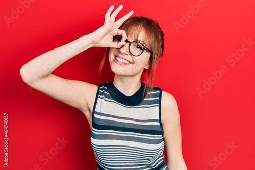 Redhead young woman wearing casual clothes and glasses smiling happy doing ok sign with hand on eye looking through fingers