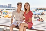 Young hispanic couple on vacation smiling happy sitting on bench at the beach