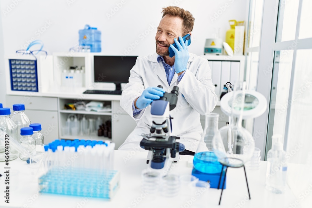 Middle age hispanic man wearing scientist uniform talking on the smartphone using microscope at laboratory Middle age hispanic man wearing scientist uniform talking on the smartphone using microscope at laboratory