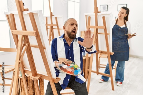 Senior artist man at art studio doing stop gesture with hands palms, angry and frustration expression