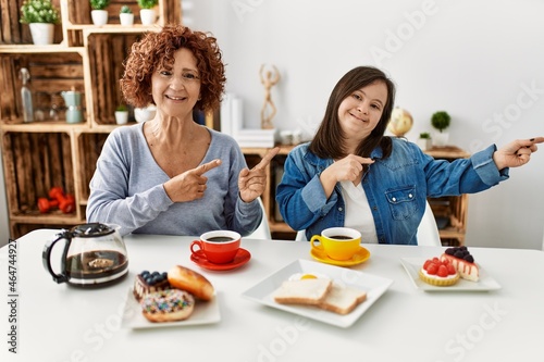 Family of mother and down syndrome daughter sitting at home eating breakfast smiling and looking at the camera pointing with two hands and fingers to the side.