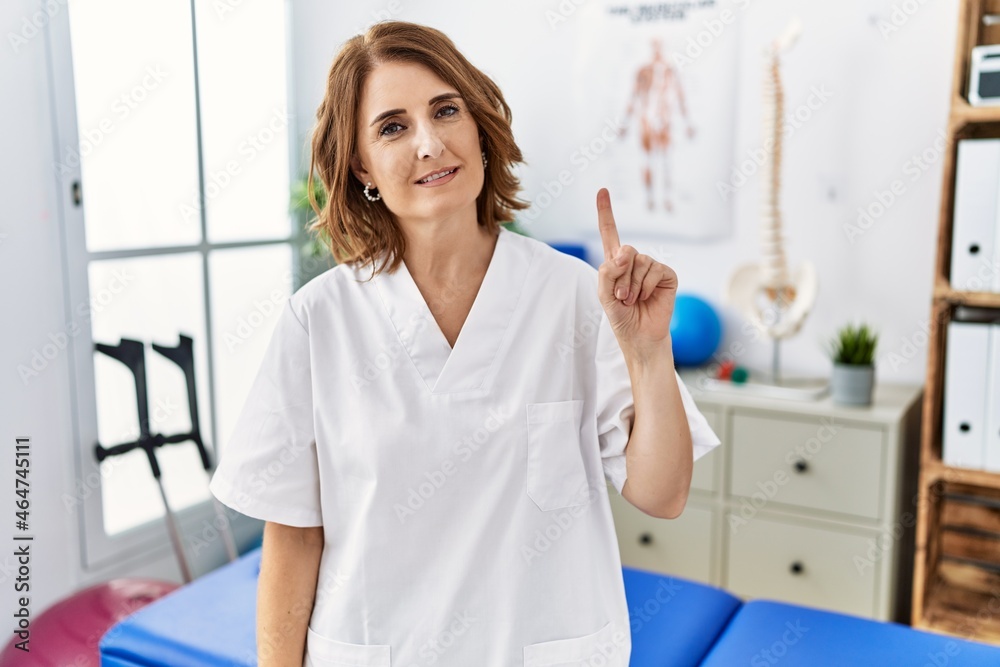 Middle age physiotherapist woman working at pain recovery clinic showing and pointing up with finger number one while smiling confident and happy. Middle age physiotherapist woman working at pain recovery clinic showing and pointing up with finger number one while smiling confident and happy.