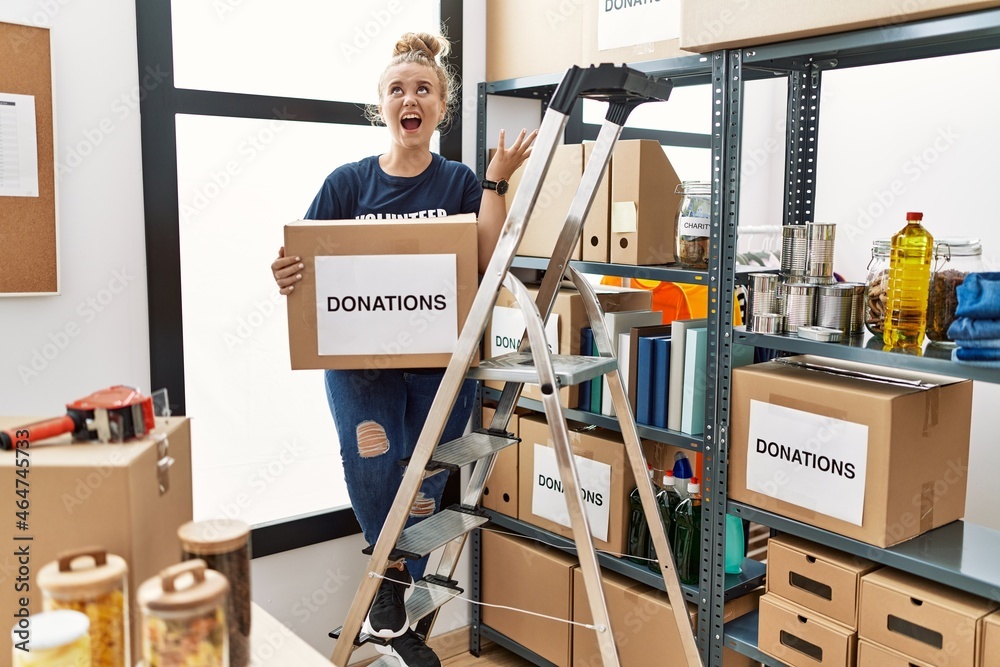 Young caucasian woman volunteer holding donations box crazy and mad shouting and yelling with aggressive expression and arms raised. frustration concept.