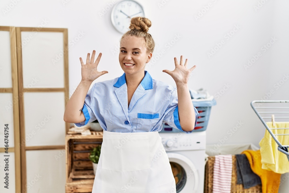 Young caucasian woman wearing cleaner uniform at the laundry room showing and pointing up with fingers number ten while smiling confident and happy.