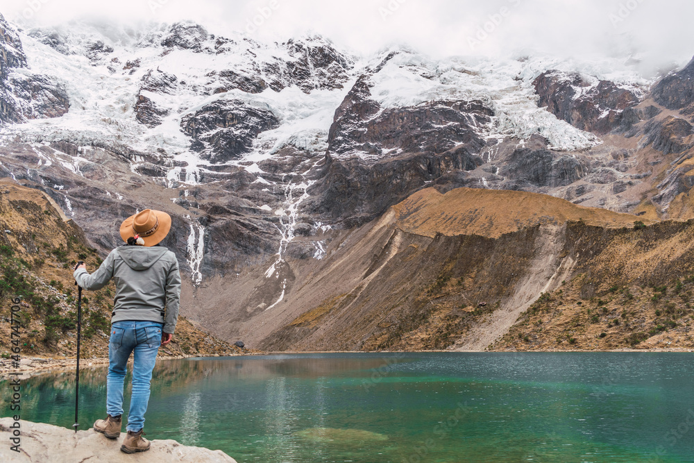 Naklejka premium hiker man with brown hat on his back standing in the humantay lagoon in the Andes mountain range of peru cusco surrounded by snow-capped mountains with clouds