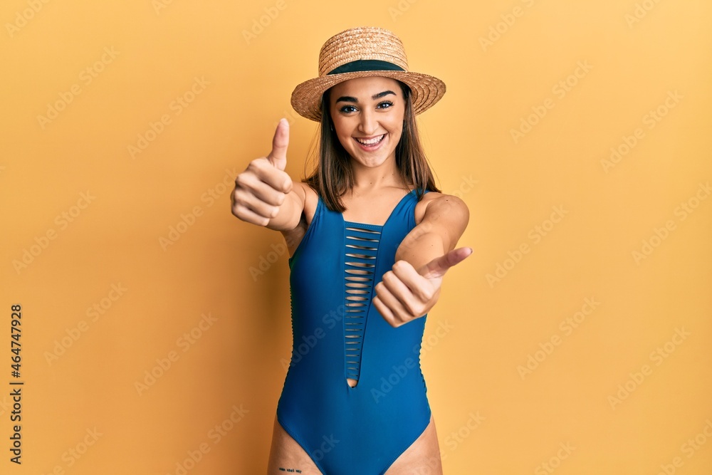 Young brunette girl wearing swimsuit and summer hat approving doing positive gesture with hand, thumbs up smiling and happy for success. winner gesture. Young brunette girl wearing swimsuit and summer hat approving doing positive gesture with hand, thumbs up smiling and happy for success. winner gesture.