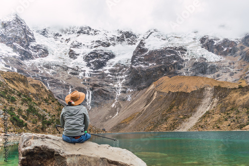 hiker man with brown hat on his back standing in the humantay lagoon in the Andes mountain range of peru cusco surrounded by snow-capped mountains with clouds