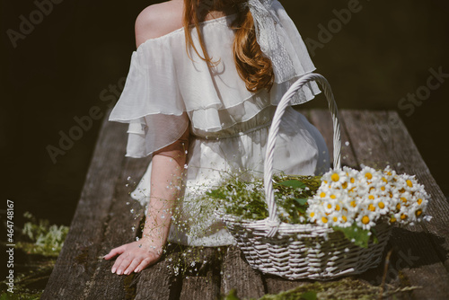 beautiful young girl with red curly hair in a long white dress sits on a wooden pier next to her stands a white straw basket with wildflowers, a rural scene on a sunny day rear view  photo