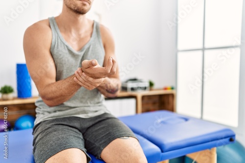 Young hispanic man suffering for hand pain sitting on massage board at clinic