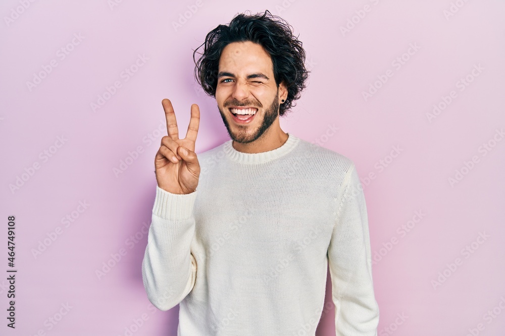 Fototapeta premium Handsome hispanic man wearing casual white sweater smiling with happy face winking at the camera doing victory sign with fingers. number two.