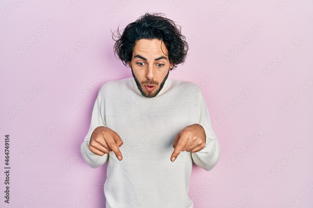 Handsome hispanic man wearing casual white sweater pointing down with fingers showing advertisement, surprised face and open mouth Handsome hispanic man wearing casual white sweater pointing down with fingers showing advertisement, surprised face and open mouth