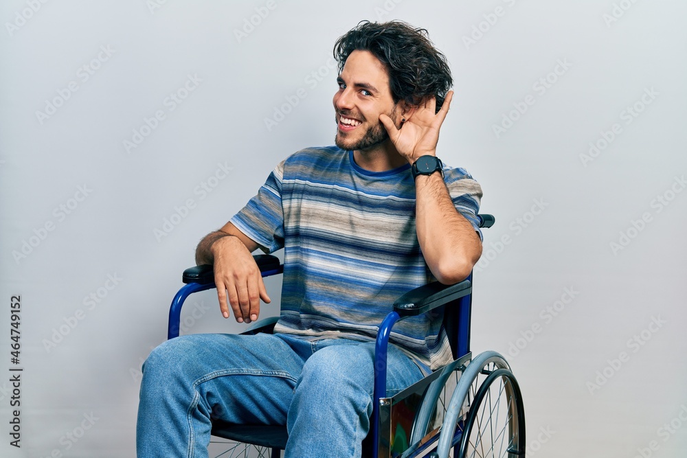 Handsome hispanic man sitting on wheelchair smiling with hand over ear listening an hearing to rumor or gossip. deafness concept.