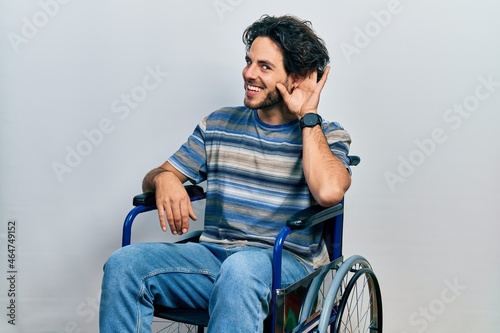 Handsome hispanic man sitting on wheelchair smiling with hand over ear listening an hearing to rumor or gossip. deafness concept.