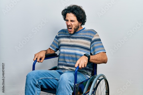 Handsome hispanic man sitting on wheelchair angry and mad screaming frustrated and furious, shouting with anger. rage and aggressive concept.