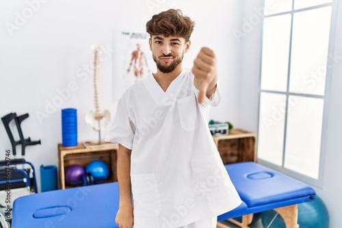 Young arab man working at pain recovery clinic looking unhappy and angry showing rejection and negative with thumbs down gesture. bad expression.