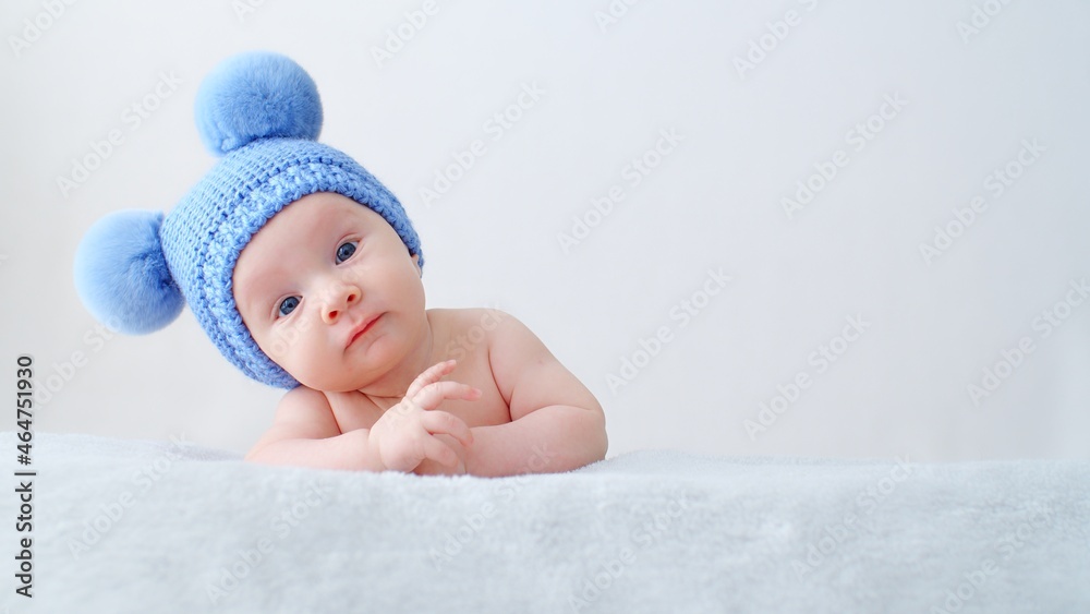 Sweet newborn baby in a blue hat with pompoms looks into the camera. Fun Beautiful, cute little infant with blue eyes looks into the camera. A calm two-month-old kid is lying on a blanket. Sweet newborn baby in a blue hat with pompoms looks into the camera. Fun Beautiful, cute little infant with blue eyes looks into the camera. A calm two-month-old kid is lying on a blanket.
