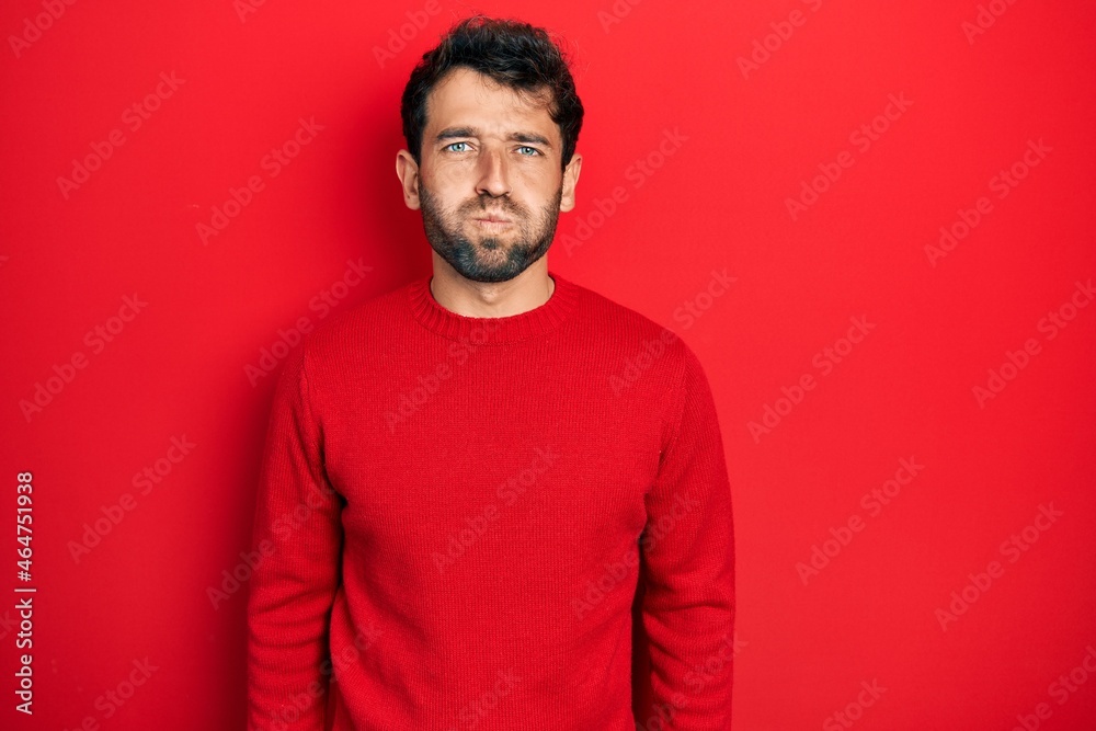 Handsome man with beard wearing casual red sweater puffing cheeks with funny face. mouth inflated with air, crazy expression. Handsome man with beard wearing casual red sweater puffing cheeks with funny face. mouth inflated with air, crazy expression.