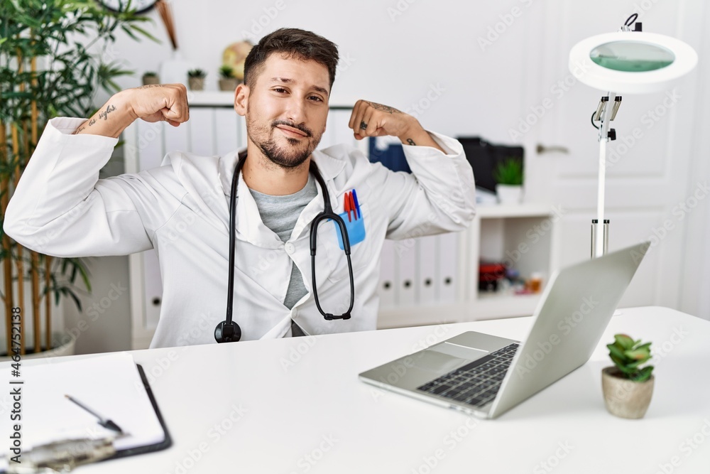 Young doctor working at the clinic using computer laptop showing arms muscles smiling proud. fitness concept. Young doctor working at the clinic using computer laptop showing arms muscles smiling proud. fitness concept.