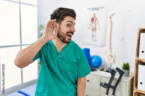 Young man with beard working at pain recovery clinic smiling with hand over ear listening an hearing to rumor or gossip. deafness concept.