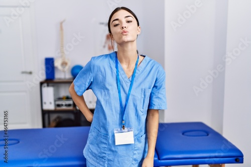 Young hispanic woman wearing physiotherapist uniform standing at clinic looking at the camera blowing a kiss on air being lovely and sexy. love expression.