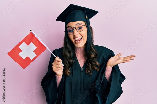Young hispanic woman wearing graduation uniform holding switzerland flag celebrating achievement with happy smile and winner expression with raised hand