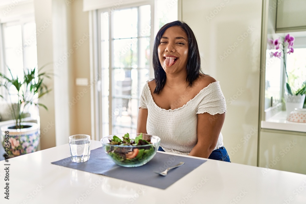 Young hispanic woman eating healthy salad at home sticking tongue out happy with funny expression. emotion concept. Young hispanic woman eating healthy salad at home sticking tongue out happy with funny expression. emotion concept.