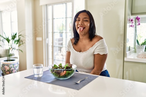 Young hispanic woman eating healthy salad at home sticking tongue out happy with funny expression. emotion concept.