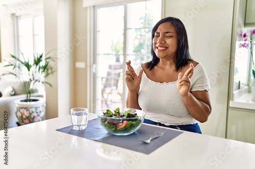 Young hispanic woman eating healthy salad at home gesturing finger crossed smiling with hope and eyes closed. luck and superstitious concept.