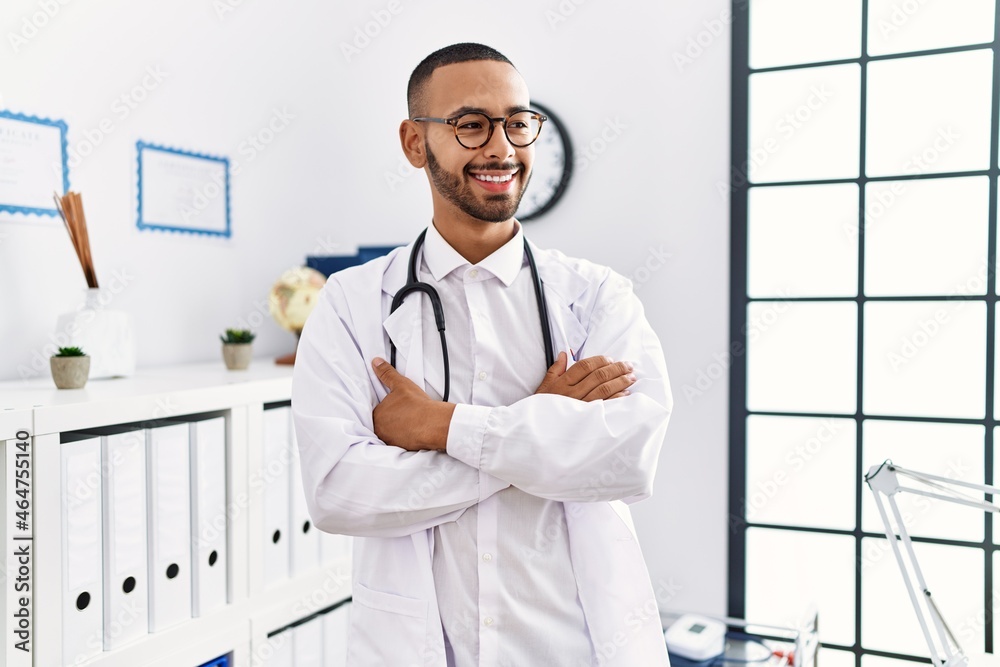Young hispanic man wearing doctor uniform with arms crossed gesture at clinic