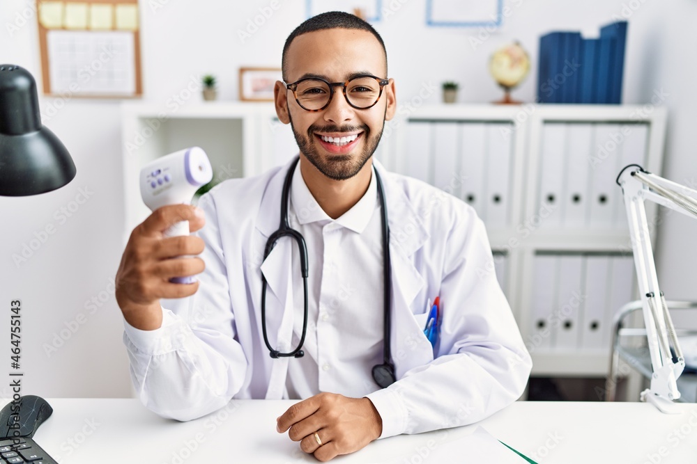 African american doctor man holding thermometer at the clinic looking positive and happy standing and smiling with a confident smile showing teeth African american doctor man holding thermometer at the clinic looking positive and happy standing and smiling with a confident smile showing teeth