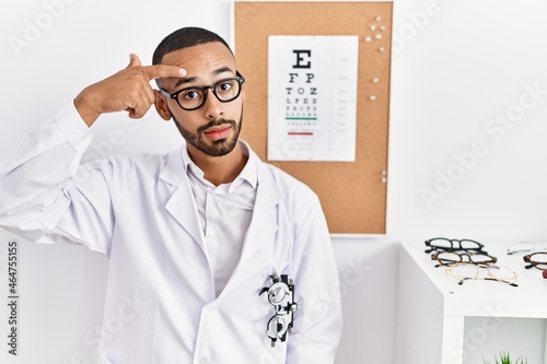 African american optician man standing by eyesight test pointing unhappy to pimple on forehead, ugly infection of blackhead. acne and skin problem photo