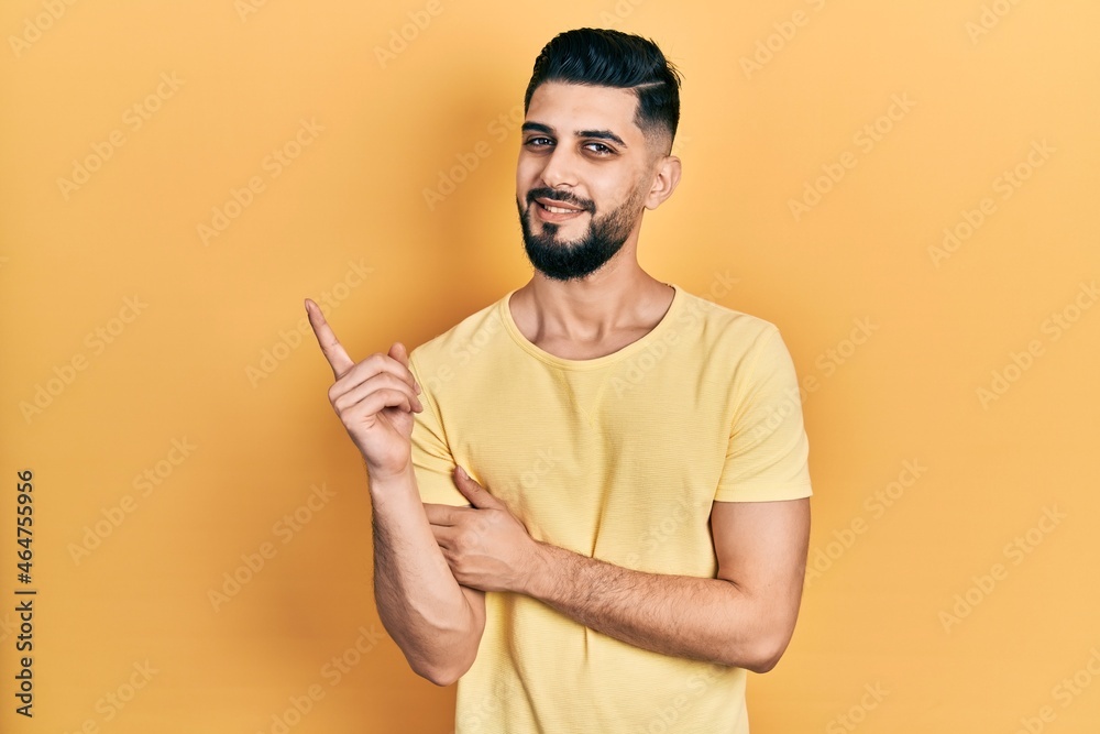 Handsome man with beard wearing casual yellow t shirt with a big smile on face, pointing with hand and finger to the side looking at the camera. Handsome man with beard wearing casual yellow t shirt with a big smile on face, pointing with hand and finger to the side looking at the camera.