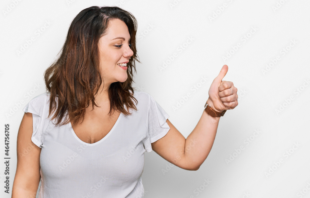 Young plus size woman wearing casual white t shirt looking proud, smiling doing thumbs up gesture to the side Young plus size woman wearing casual white t shirt looking proud, smiling doing thumbs up gesture to the side