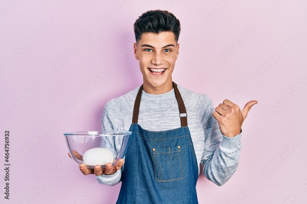 Young hispanic man wearing apron holding bowl of bread dough pointing thumb up to the side smiling happy with open mouth Young hispanic man wearing apron holding bowl of bread dough pointing thumb up to the side smiling happy with open mouth