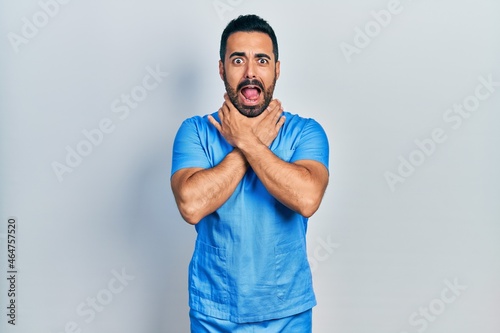 Handsome hispanic man with beard wearing blue male nurse uniform shouting and suffocate because painful strangle. health problem. asphyxiate and suicide concept.