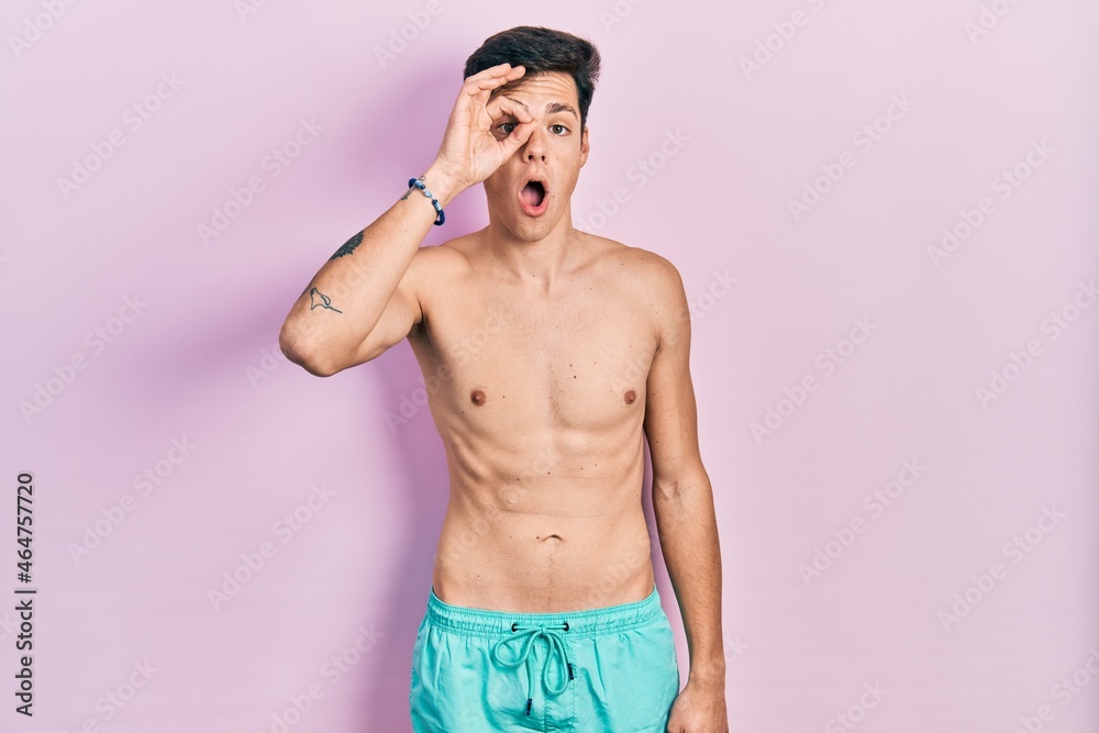 Fototapeta premium Young hispanic man wearing swimwear shirtless doing ok gesture shocked with surprised face, eye looking through fingers. unbelieving expression.