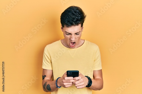 Young hispanic man using smartphone typing message angry and mad screaming frustrated and furious, shouting with anger. rage and aggressive concept. © Krakenimages.com