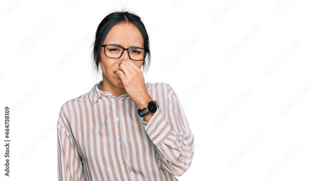 Young hispanic girl wearing casual clothes and glasses smelling something stinky and disgusting, intolerable smell, holding breath with fingers on nose. bad smell Young hispanic girl wearing casual clothes and glasses smelling something stinky and disgusting, intolerable smell, holding breath with fingers on nose. bad smell