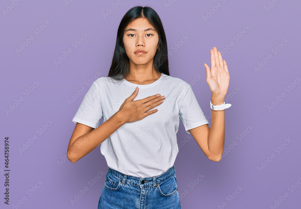 Beautiful young asian woman wearing casual white t shirt swearing with hand on chest and open palm, making a loyalty promise oath Beautiful young asian woman wearing casual white t shirt swearing with hand on chest and open palm, making a loyalty promise oath