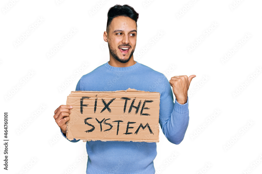 Young arab man holding fix the system banner cardboard pointing thumb up to the side smiling happy with open mouth Young arab man holding fix the system banner cardboard pointing thumb up to the side smiling happy with open mouth
